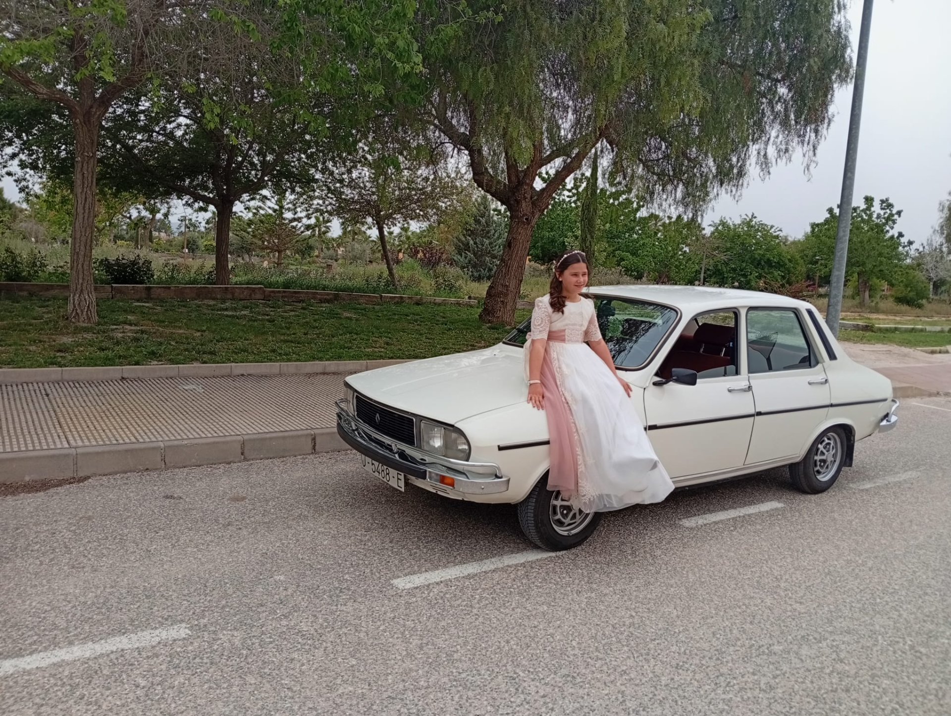En esta foto apreciamos una chica con un bonito y elegante vestido largo de comunión. La chica se encuentra sentada en la parte delantera del coche. 